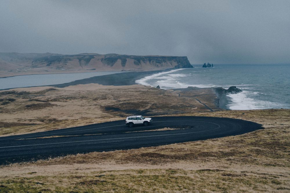 White SUV driving along a winding coastal road near Reynisfjara beach on Iceland’s South Coast